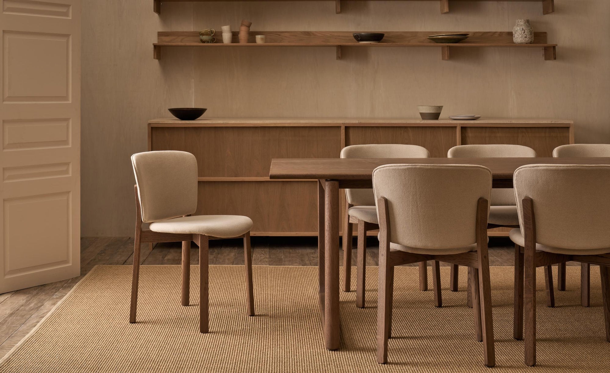 A minimalist dining room features Danish lines with the Wooden Stories Hover Dining Table in Espresso Oak, six beige cushioned chairs, a wooden sideboard, and wall-mounted shelves for pottery. Neutral tones and a woven rug complete the look.