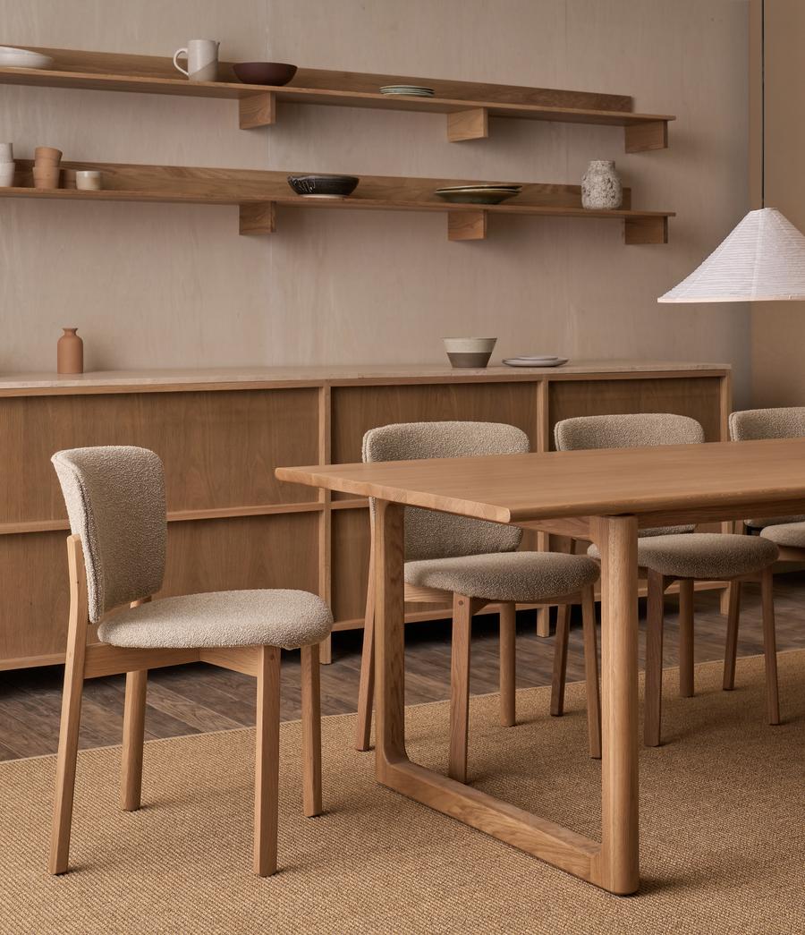 A minimalist dining room features Danish lines with a Wooden Stories Hover Dining Table in Light Oak, six upholstered chairs, wall-mounted wooden shelves displaying ceramics, and a pendant lamp—all in neutral tones and natural wood finishes.