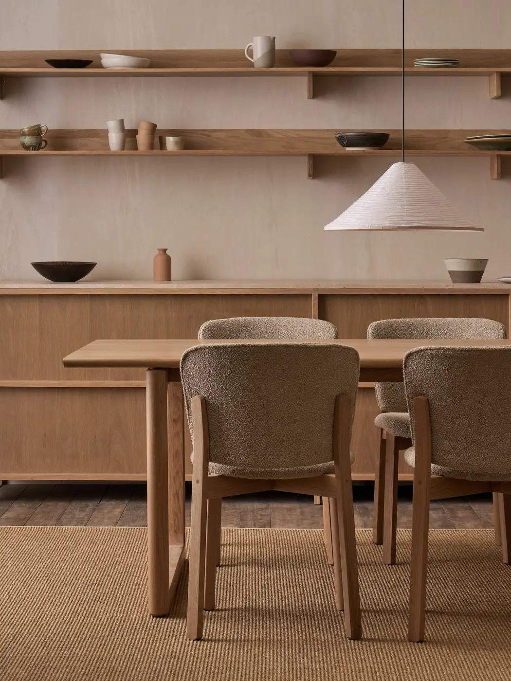 A minimalist dining room with Danish lines features the Hover Dining Table in Light Oak by Wooden Stories, four upholstered chairs, open shelves with ceramics, a pendant lamp, and neutral-toned furniture and decor.