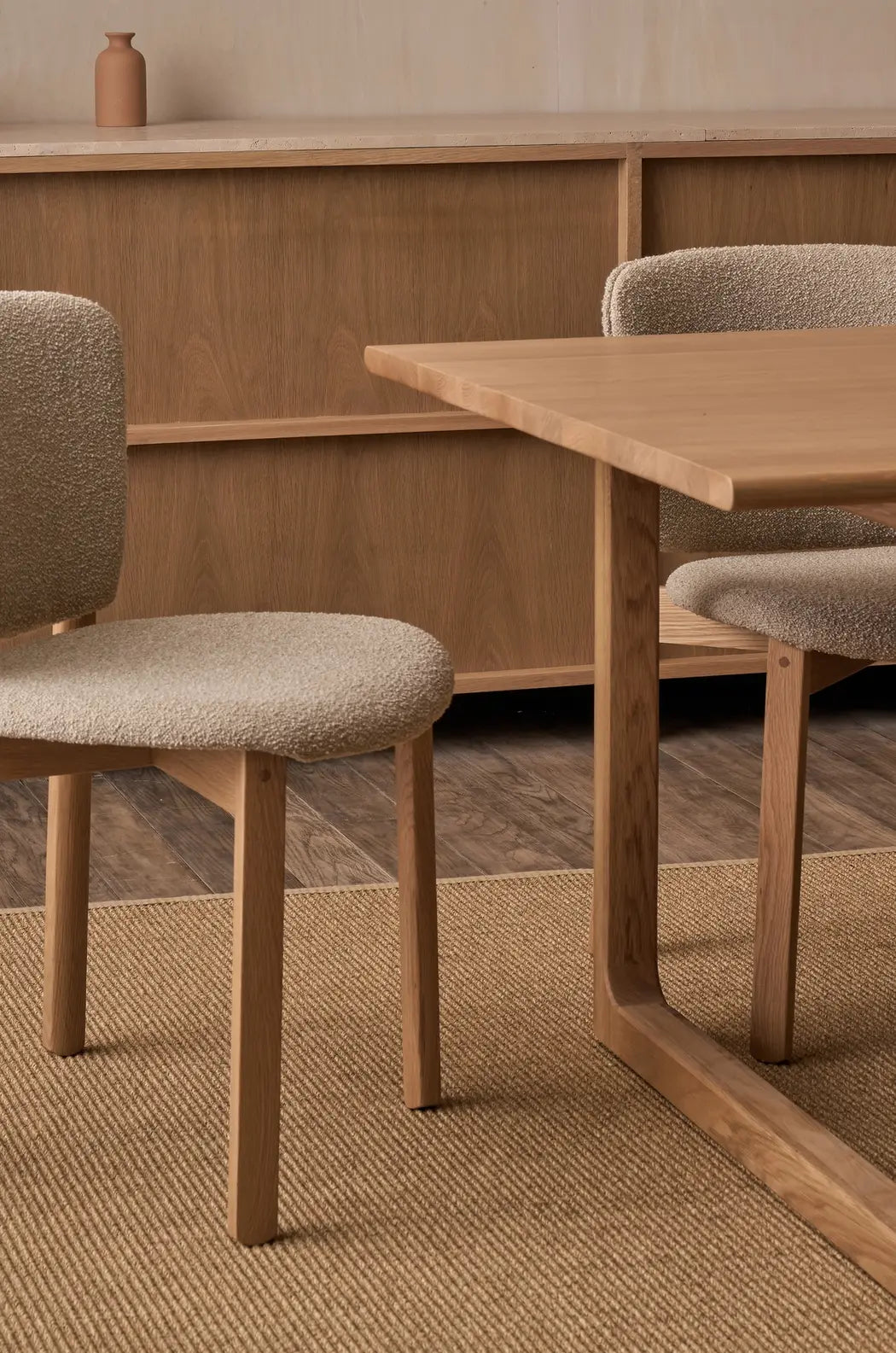A modern dining room featuring the Hover Dining Table in Light Oak by Wooden Stories, two beige upholstered chairs, a Danish-style wooden sideboard with a small vase on top, and a textured beige rug over the wooden floor.