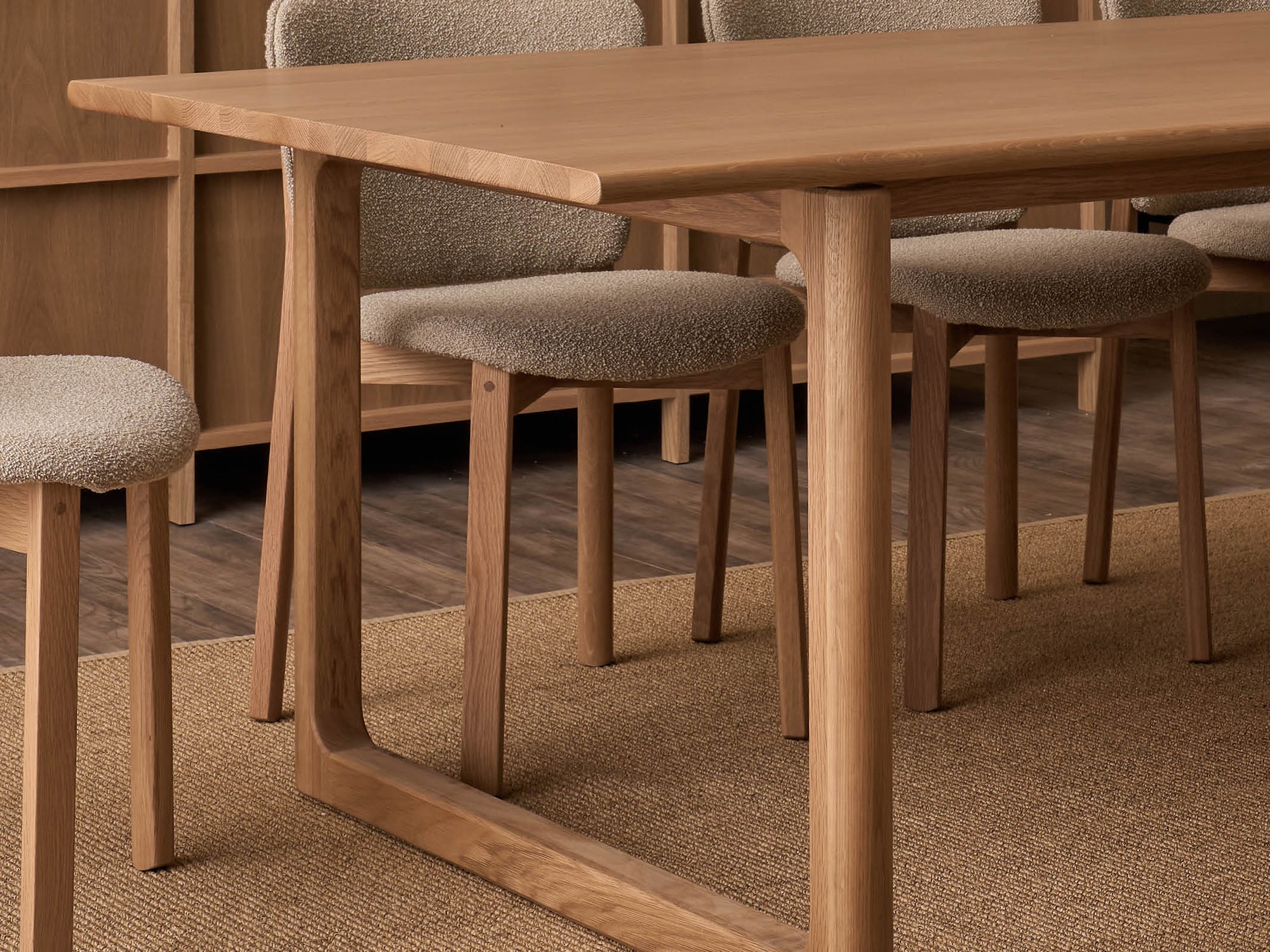 A close-up of the Wooden Stories Hover Dining Table in Light Oak with four cushioned chairs on a woven rug, all in neutral tones, set atop a dark wooden dining room floor.