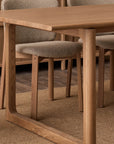 A close-up of the Wooden Stories Hover Dining Table in Light Oak with four cushioned chairs on a woven rug, all in neutral tones, set atop a dark wooden dining room floor.