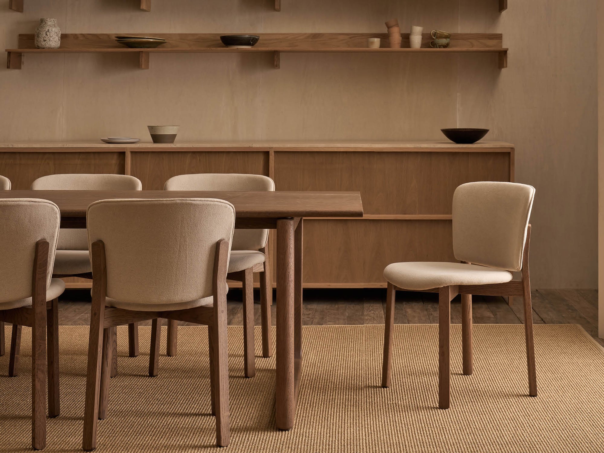 A dining room featuring Wooden Stories’ Hover Dining Table in Espresso Oak, six beige cushioned chairs, a light tan rug, minimalist pottery on wooden shelves, and a wooden sideboard creates a modern, neutral-toned aesthetic.