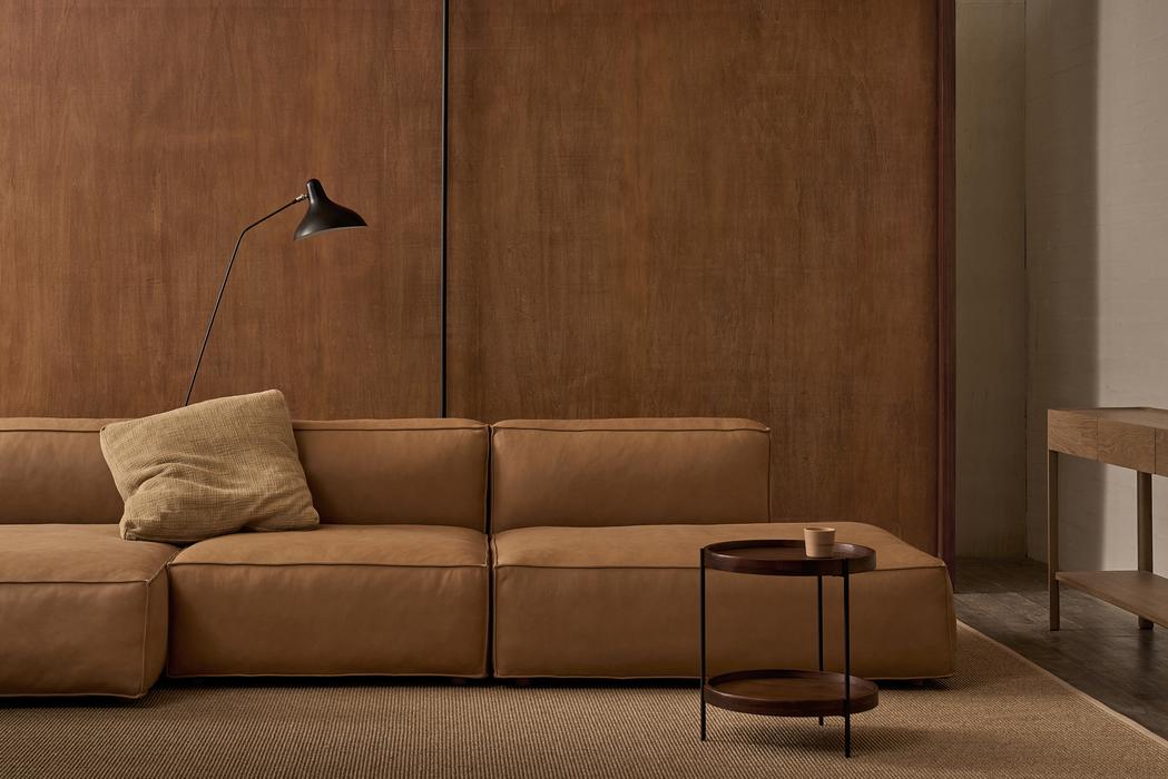 A modern living room featuring a tan leather sectional, beige cushion, black floor lamp, Wooden Stories Humla Round Side Table, and wooden paneled walls in a minimalist, neutral-toned Danish design.