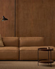 A modern living room featuring a tan leather sectional, beige cushion, black floor lamp, Wooden Stories Humla Round Side Table, and wooden paneled walls in a minimalist, neutral-toned Danish design.