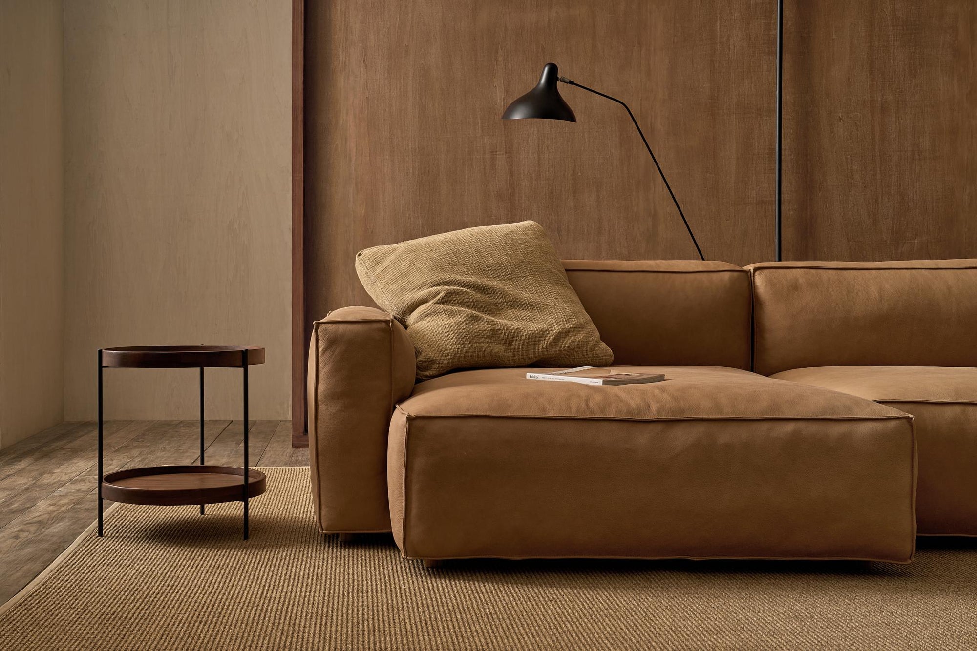 A modern living room features a tan leather sofa, a large beige pillow, a black floor lamp, and the Humla Round Side Table by Wooden Stories. A textured beige rug covers the wooden floor, complemented by wooden panels in the background.