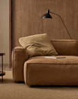A modern living room features a tan leather sofa, a large beige pillow, a black floor lamp, and the Humla Round Side Table by Wooden Stories. A textured beige rug covers the wooden floor, complemented by wooden panels in the background.