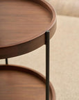 Close-up of the Humla Round Side Table by Wooden Stories, a Danish-inspired design with a raised edge, metal legs, and two shelves, shown on a light-colored textured rug—evoking classic Hans Bolling tray tables.