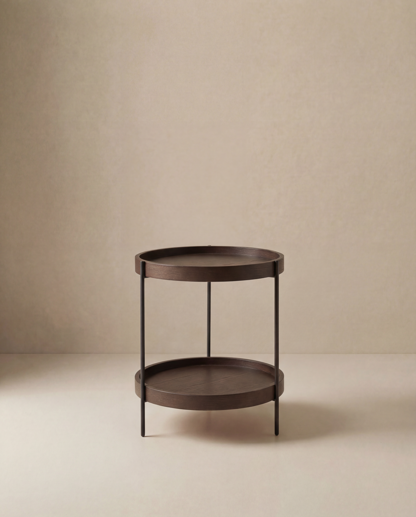 The Humla End Table by Wooden Stories features a round, dark wood design with two tiers and slim metal legs, showcased against a beige minimalist background.