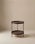 The Humla End Table by Wooden Stories features a round, dark wood design with two tiers and slim metal legs, showcased against a beige minimalist background.