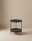The Wooden Stories Humla Round Side Table features a minimalist black metal design with two tiers and four slender legs, exemplifying refined Danish style against a plain beige background.