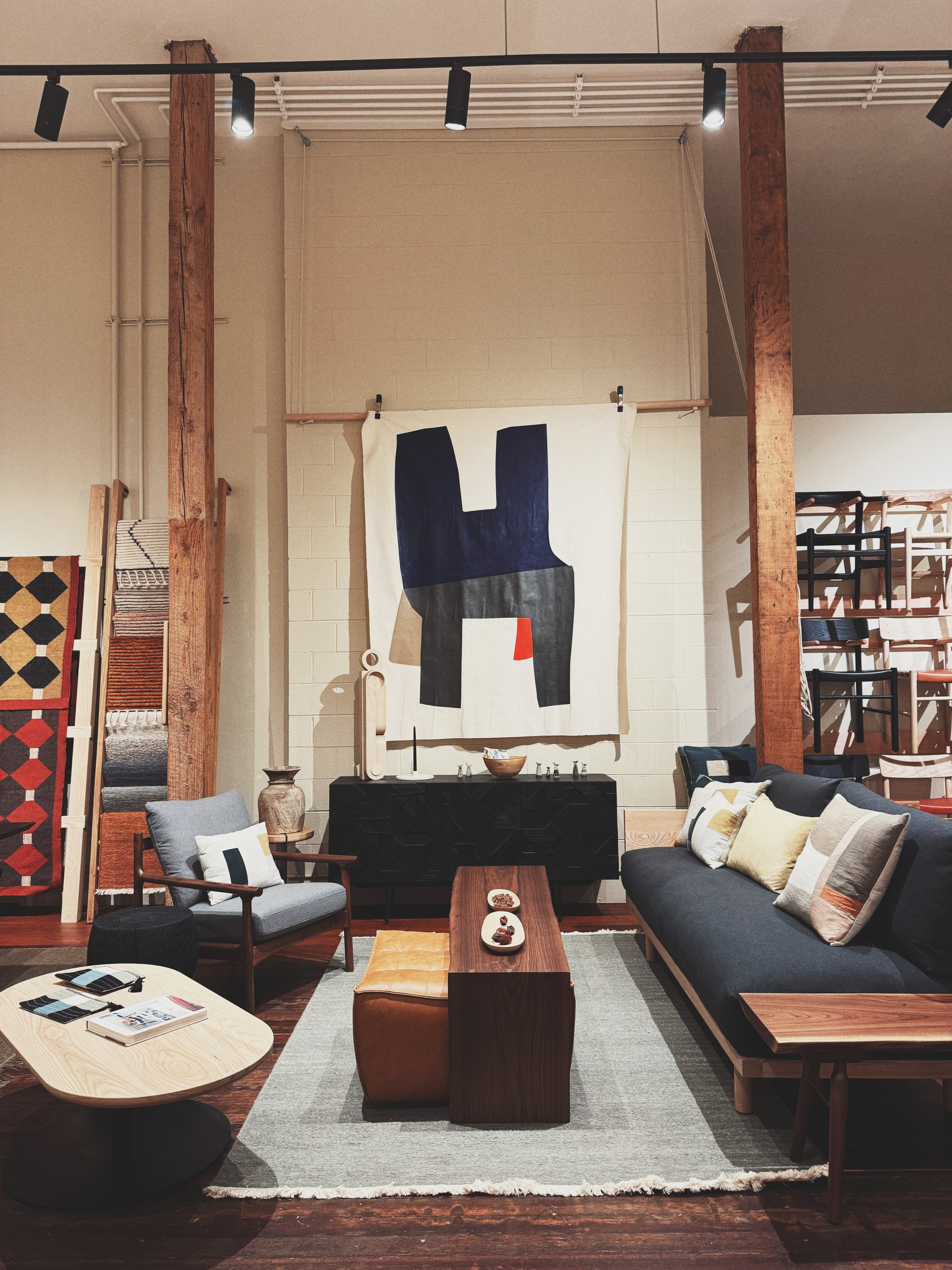 A modern and inviting showroom setup with a dark blue sofa, a light wood coffee table, and accent furniture. Decorative pillows and a geometric wall tapestry add color, while a black credenza and art pieces line the back wall. 