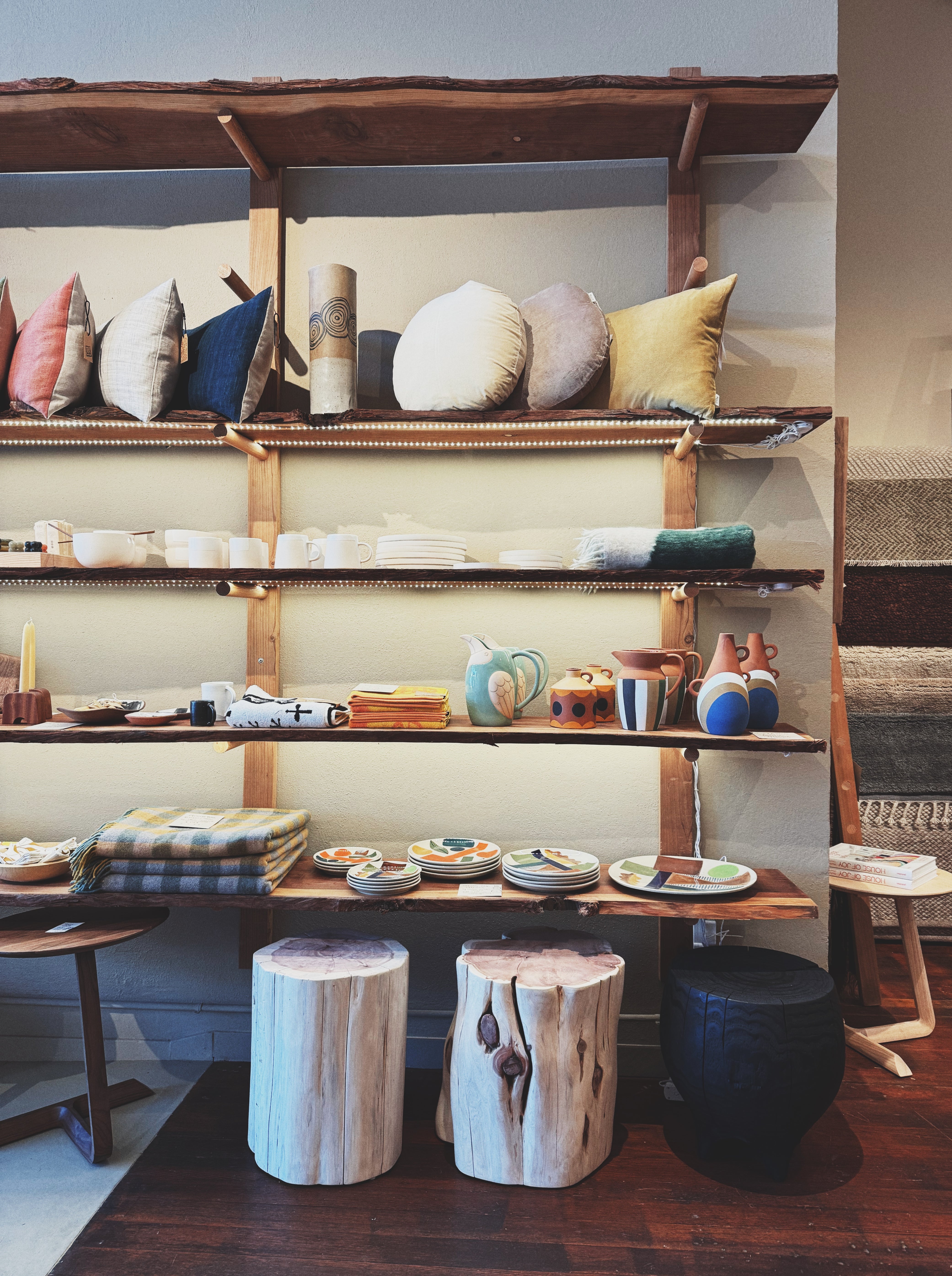 A cozy retail display featuring a wooden shelving unit filled with home goods. The top shelf holds decorative throw pillows in various colors, while lower shelves display ceramics, mugs, pitchers, textiles, and folded blankets. 