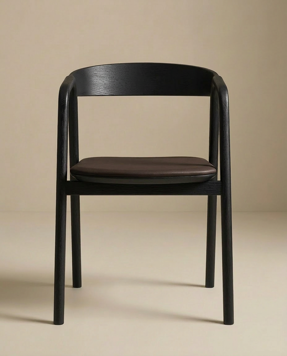 The Inlay Dining Chair | Black Oak and Leather by Wooden Stories features a curved backrest, brown leather seat, and solid black oak frame, set against a neutral beige backdrop. Part of the Danish Collection for timeless elegance.