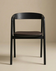 The Inlay Dining Chair | Black Oak and Leather by Wooden Stories features a curved backrest, brown leather seat, and solid black oak frame, set against a neutral beige backdrop. Part of the Danish Collection for timeless elegance.