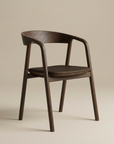 The Inlay Dining Chair by Wooden Stories features espresso oak, a leather cushioned seat, curved backrest, and armrests, shown against a plain beige background.