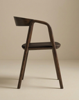 The Inlay Dining Chair by Wooden Stories features espresso oak, leather accents, and sculptural curves. Part of the Danish Collection, it is shown in profile against a beige background with a curved backrest and seat.
