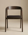 The Inlay Dining Chair by Wooden Stories features an espresso oak frame, a curved backrest, and a black leather seat. Photographed against a beige background, it’s part of the brand’s Danish Collection.