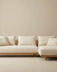 The Islet Sofa | Fabric by Wooden Stories is a modern, minimalist cream-colored sectional with wooden legs and four matching cushions, placed against a beige wall and floor.