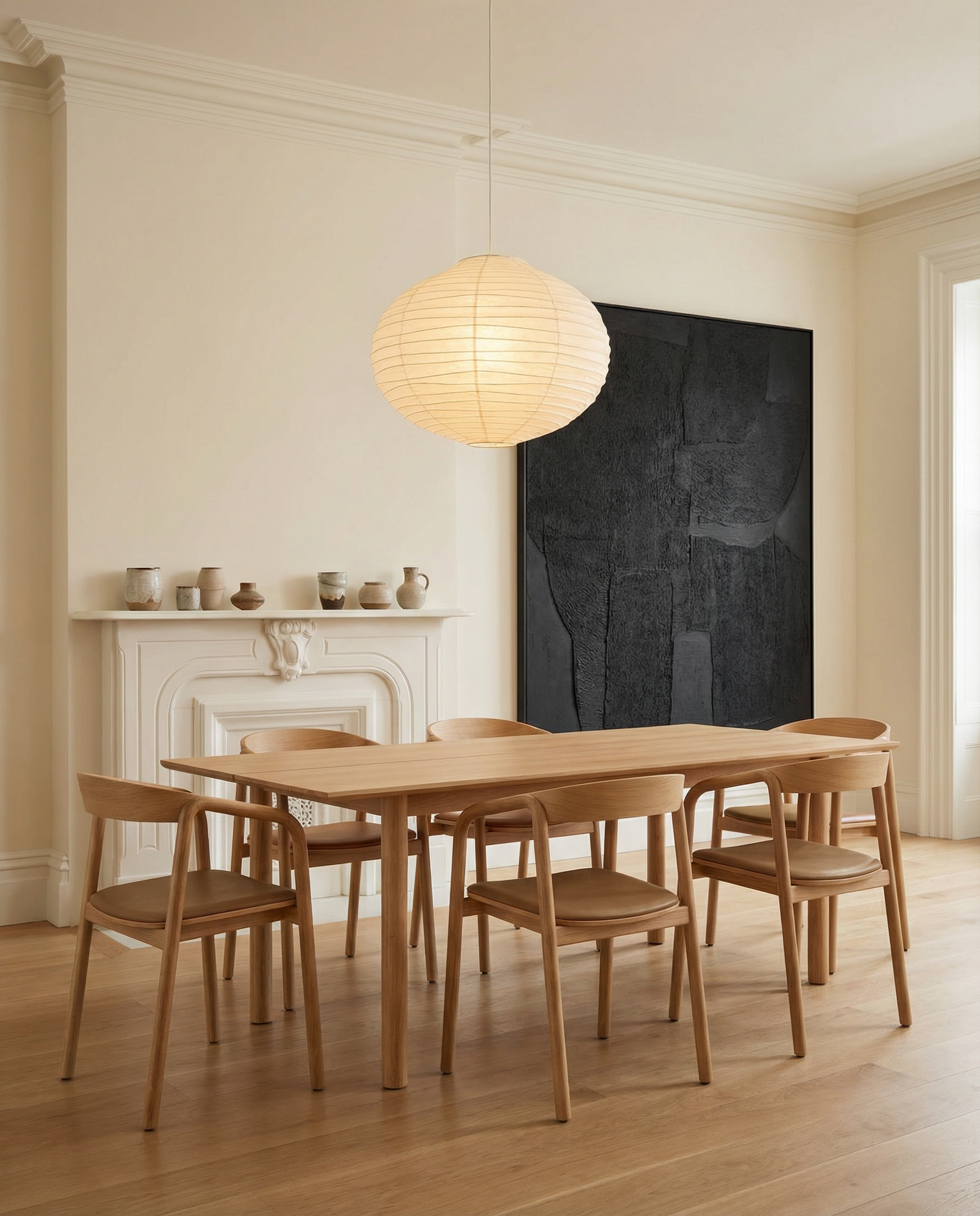 A minimalist dining room features a wooden table with six Inlay Dining Chairs in Light Oak and Leather by Wooden Stories, a white fireplace adorned with pottery, a black abstract painting, and a round paper lantern. Pale walls and wood floors add warmth.