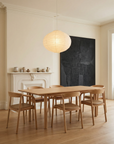 A minimalist dining room features a wooden table with six Inlay Dining Chairs in Light Oak and Leather by Wooden Stories, a white fireplace adorned with pottery, a black abstract painting, and a round paper lantern. Pale walls and wood floors add warmth.
