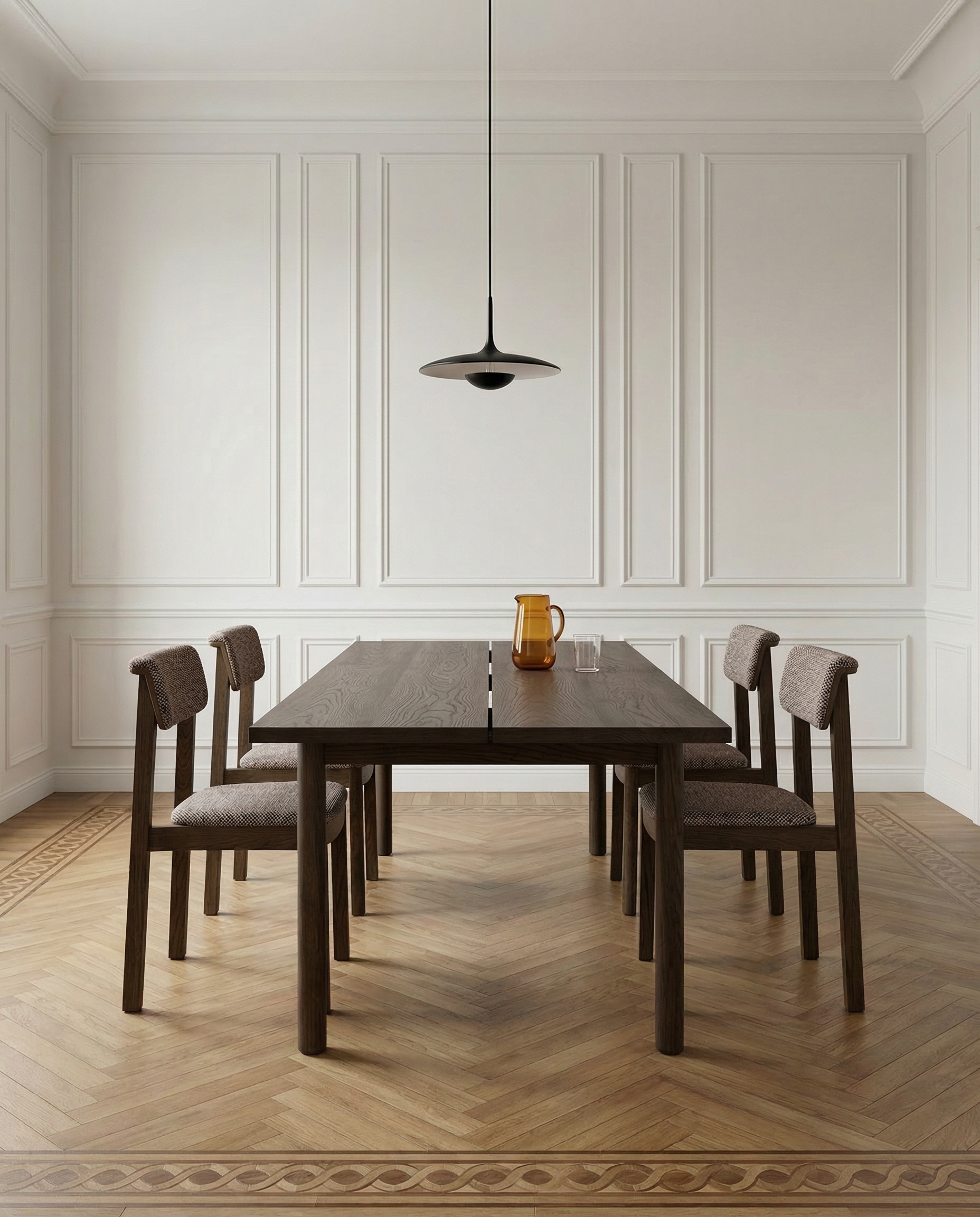 Minimalist dining room with a dark wooden table, four Wooden Stories Noki Dining Chairs in Espresso Oak with Fabric, a glass and amber pitcher on the table, black pendant light overhead, and white paneled walls with wood flooring.