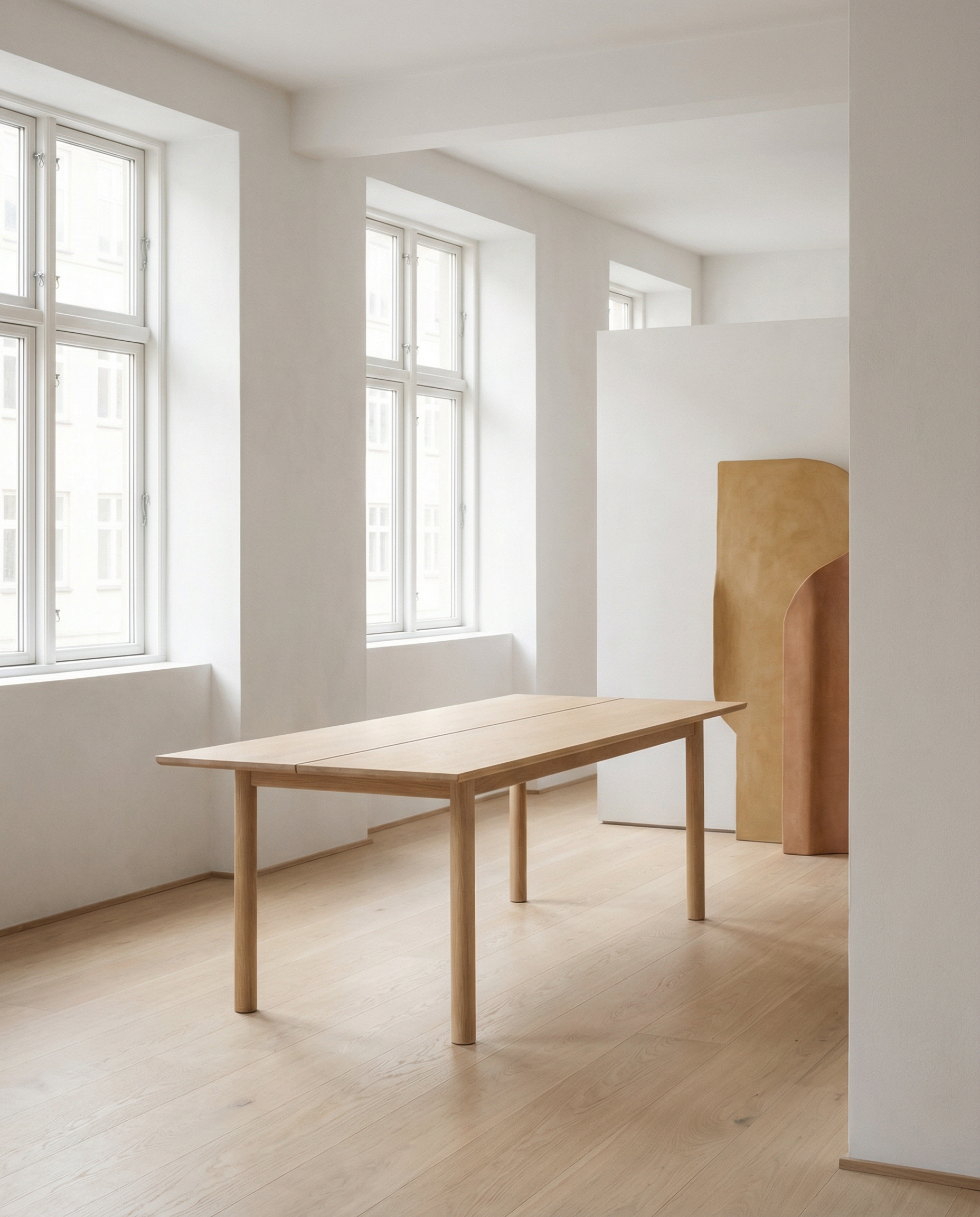 A minimalist room with light wood floors and white walls showcases the Wooden Stories Itamae Dining Table in light oak. Large windows brighten the space, while abstract beige sculptures lend elegant style to the uncluttered interior.