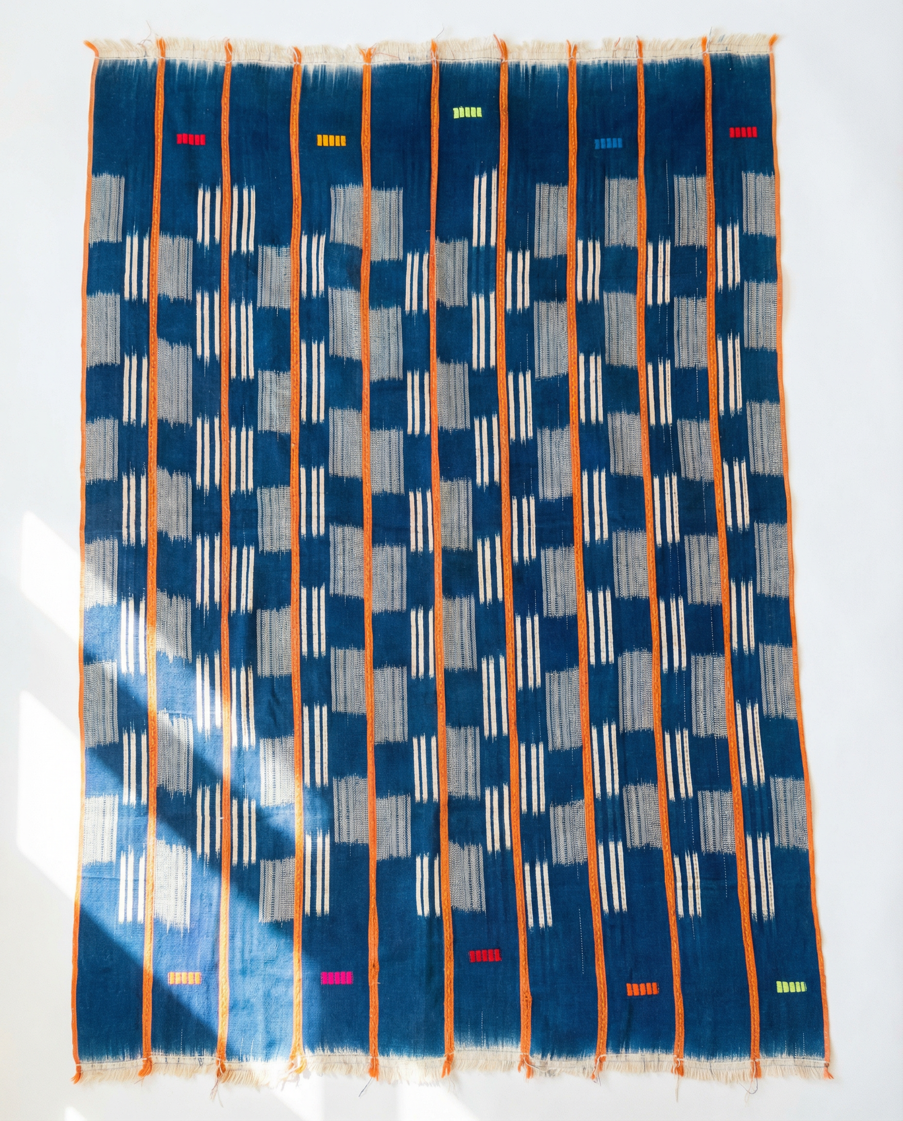The Indigo Horizon — Vintage Baule Cloth Throw by Riad Jardin Des Biehn is a handwoven cotton textile featuring vertical orange stripes, blue and gray checkered patterns, and green, red, and yellow bars on an indigo background.