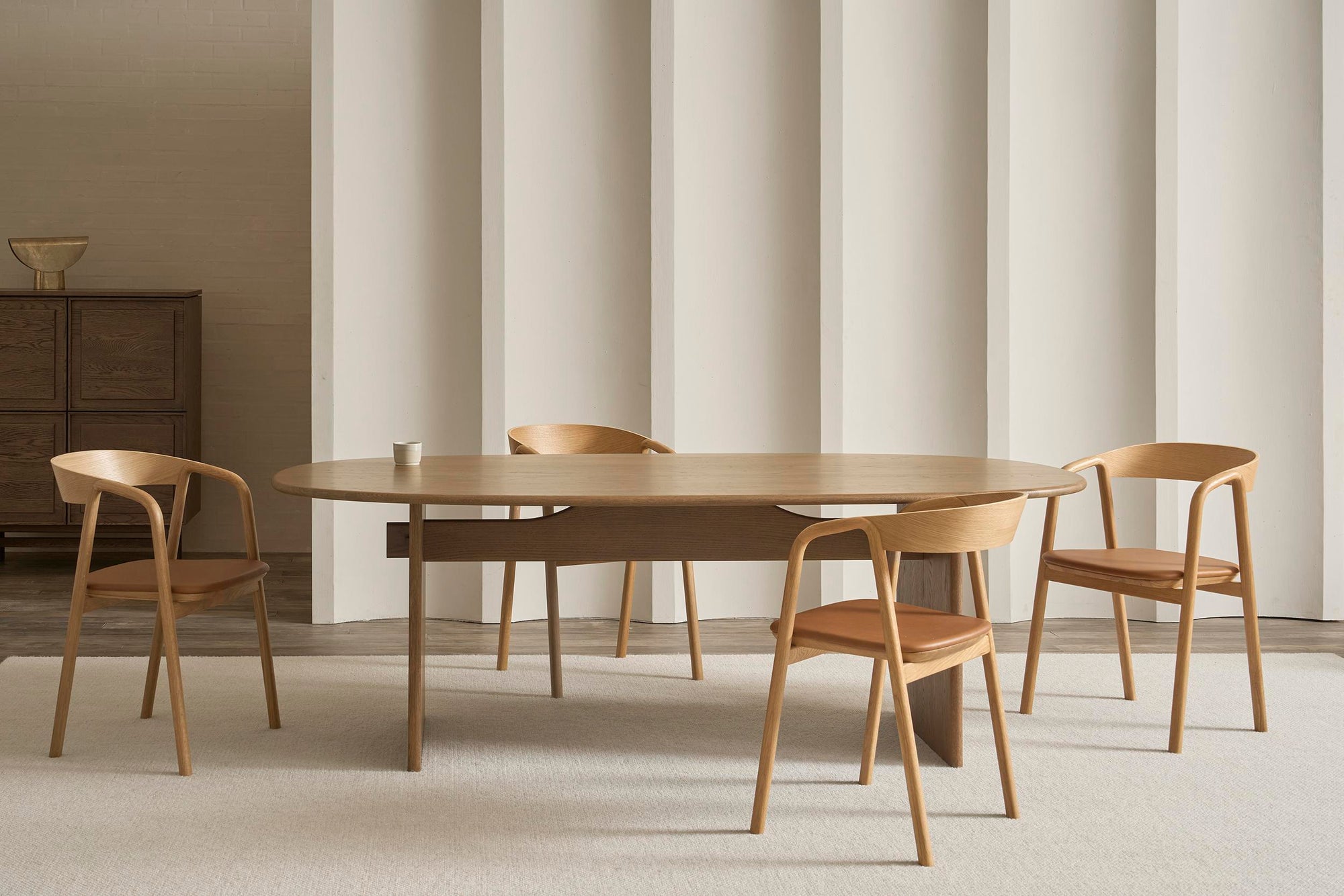 A modern dining room with a wooden oval table and four Wooden Stories Inlay Dining Chairs in light oak and leather, featuring plywood backrests and upholstered seats, a single cup on the table, light beige vertically lined walls, and a carpeted floor.