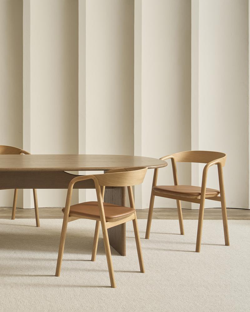 A minimalist dining room with a wooden oval table and two Wooden Stories Inlay Dining Chairs in light oak and leather sits on a light beige carpet, accented by a cream wall with vertical decorative panels.