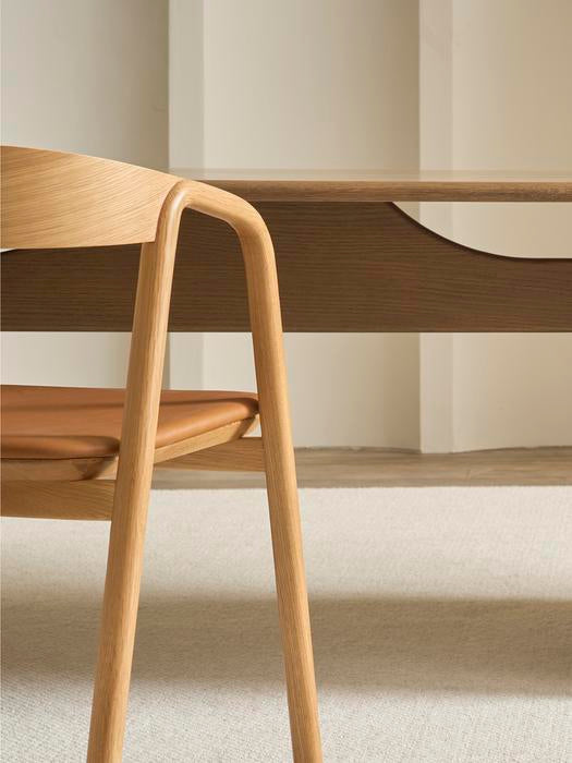 A close-up of the Wooden Stories Inlay Dining Chair in light oak and leather, shown beside a matching wooden table on a beige carpet against a plain white wall.