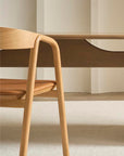 A close-up of the Wooden Stories Inlay Dining Chair in light oak and leather, shown beside a matching wooden table on a beige carpet against a plain white wall.