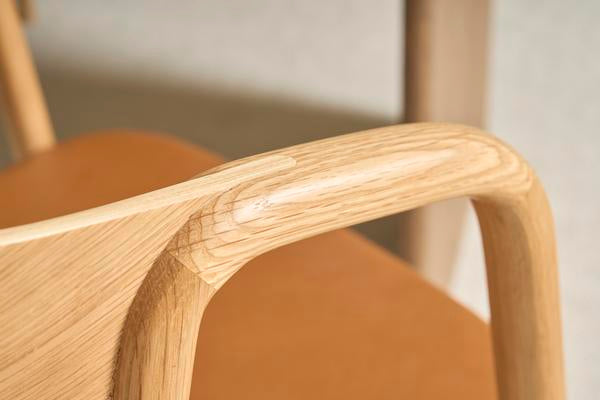 Close-up of the Wooden Stories Inlay Dining Chair in Light Oak and Leather, showing its smooth, rounded light wood armrest and warm brown leather seat; another chair is softly blurred in the background.