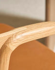 Close-up of the Wooden Stories Inlay Dining Chair in Light Oak and Leather, showing its smooth, rounded light wood armrest and warm brown leather seat; another chair is softly blurred in the background.