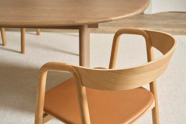 The Wooden Stories Inlay Dining Chair in Light Oak and Leather features a curved plywood backrest and tan leather seat, shown beside a round wooden table on a light rug.