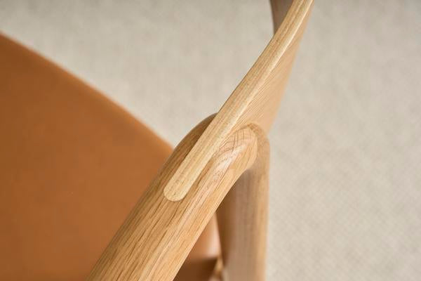 Close-up of Wooden Stories’ Inlay Dining Chair in Light Oak and Leather, showcasing the armrest and plywood backrest joint, smooth wood grain, and tan leather upholstered seat against a softly blurred neutral background.