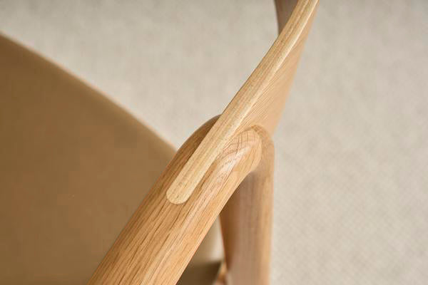 Close-up of the Wooden Stories Inlay Dining Chair in Light Oak and Leather. The image highlights its smooth solid oak armrest, seamless craftsmanship, and neutral leather seat from our Danish Collection, set against a softly blurred background.