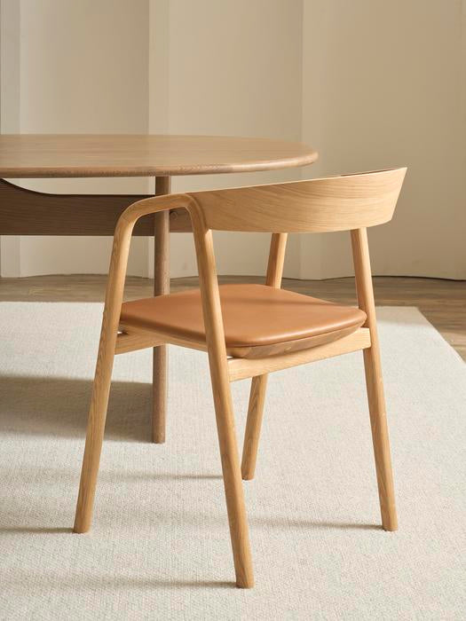 The Wooden Stories Inlay Dining Chair in light oak and leather features a curved plywood backrest and tan seat, positioned next to a round wooden table on a light rug in a minimalist room.