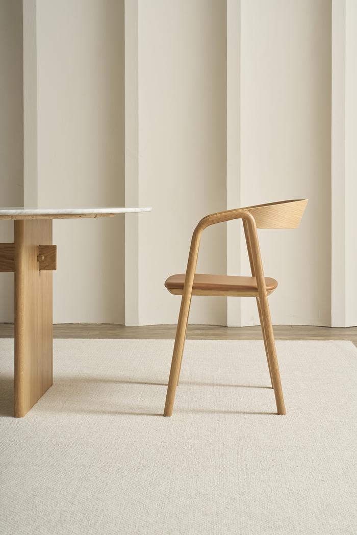 The Wooden Stories Inlay Dining Chair in light oak and leather sits beside a wooden table with a white top, placed on a light rug against a neutral, vertically paneled wall.