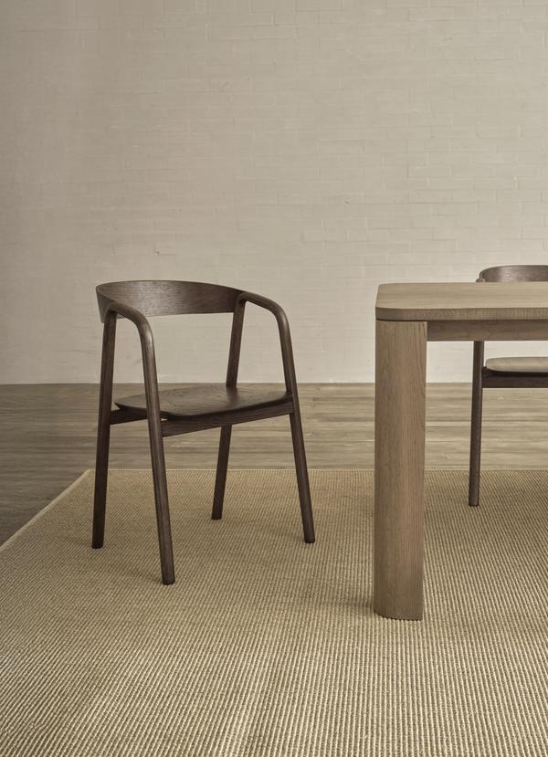 The Wooden Stories Inlay Dining Chair | Espresso Oak, featuring a curved plywood backrest, sits by a wooden table on a beige textured rug in a minimalist room with light brick walls.