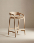 The Inlay Counter Stool | Wood Seat by Wooden Stories features a modern Danish design with a curved backrest, armrests, and footrest in smooth light wood—ideal for stylish kitchen island seating against a neutral beige background.