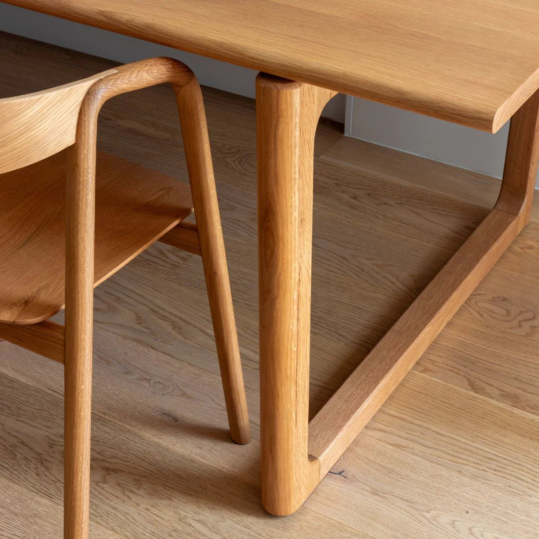 A close-up of the Wooden Stories Inlay Dining Chair in Light Oak, featuring a smooth plywood backrest, solid wood arms, and rounded edges, paired with a matching table on a natural wood floor that highlights its modern design and visible grain.