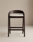 The Inlay Counter Stool | Fabric Seat by Wooden Stories features a modern solid oak frame, curved backrest, and light cushioned seat, presented against a neutral beige background.