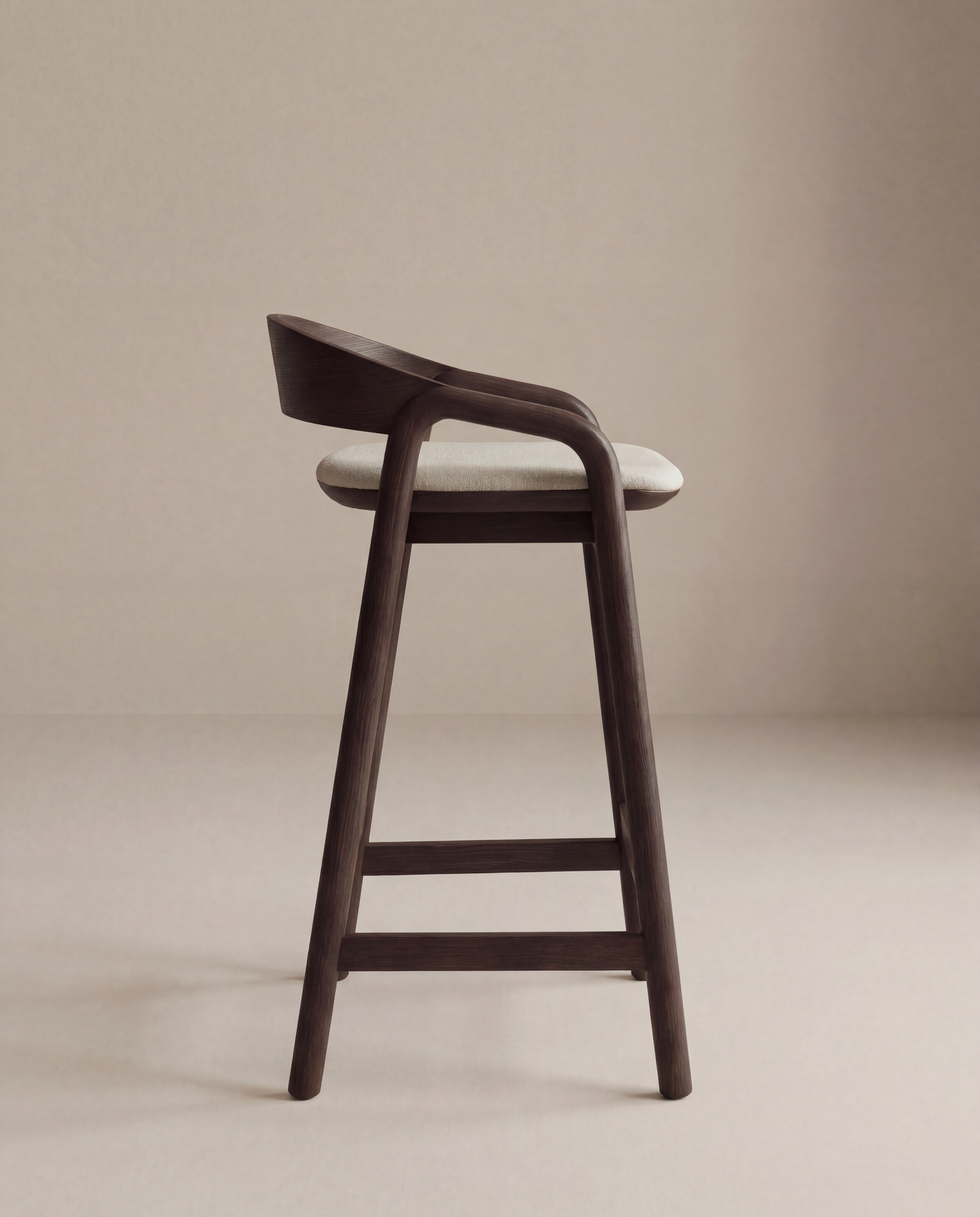 The Inlay Counter Stool | Fabric Seat by Wooden Stories features a modern dark solid oak frame, a curved backrest, and a light cushioned seat, photographed against a plain beige background.