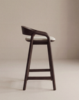 The Inlay Counter Stool | Fabric Seat by Wooden Stories features a modern dark solid oak frame, a curved backrest, and a light cushioned seat, photographed against a plain beige background.