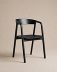 A sleek black "Inlay Dining Chair | Wood" by Wooden Stories, featuring a curved backrest and armrests inspired by the Danish Collection, is showcased against a plain beige background.