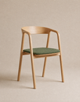 An Inlay Dining Chair | Fabric by Wooden Stories, crafted in solid oak with armrests, a curved backrest, and a green cushioned seat, is placed against a neutral beige background.