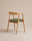 A modern solid oak wooden chair with a curved backrest and green upholstered seat, positioned at an angle against a neutral beige background, from Wooden Stories’ Inlay Dining Chair | Fabric collection.