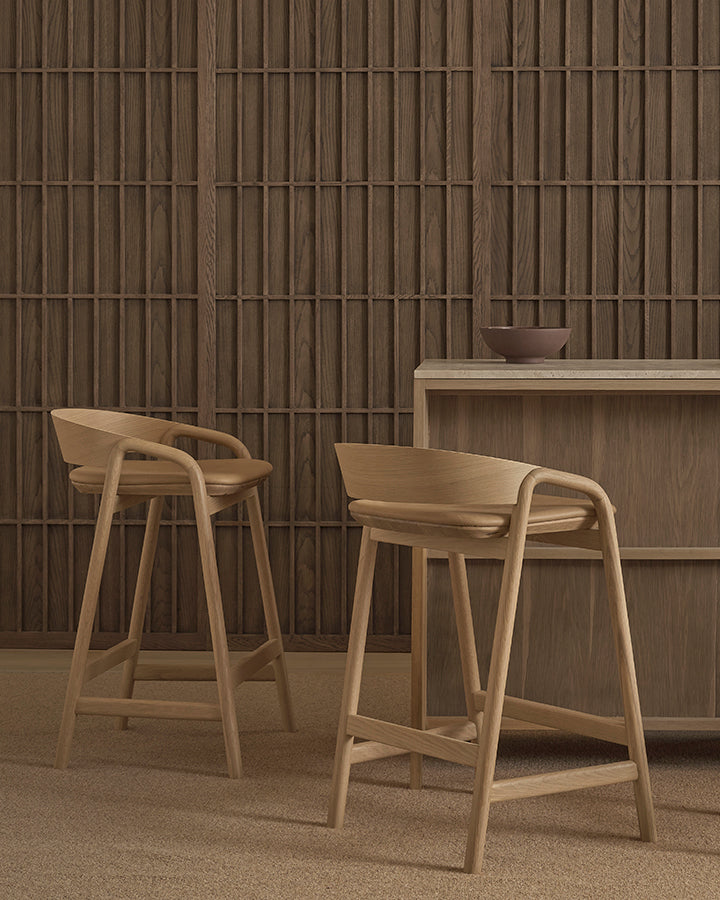 Two Wooden Stories Inlay Counter Stools with leather seats and curved backs sit by a wood counter; a ceramic bowl rests on the minimalist, modern Danish Collection-inspired setting with a textured panel wall.