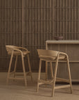Two Wooden Stories Inlay Counter Stools with leather seats and curved backs sit by a wood counter; a ceramic bowl rests on the minimalist, modern Danish Collection-inspired setting with a textured panel wall.